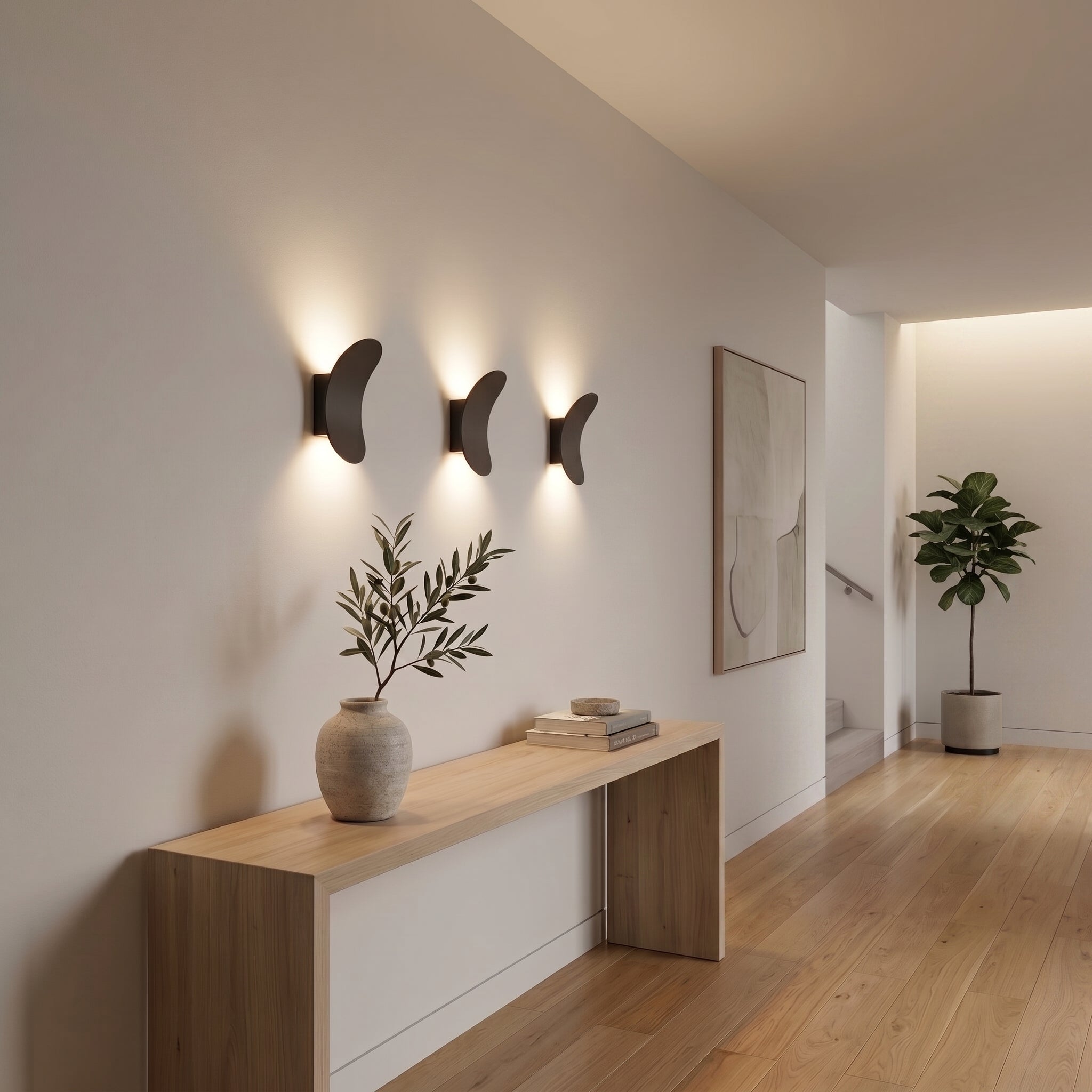 Three contemporary, dark curved back-lit LED wall sconces mounted in a row above a styled light wood console table in a minimal hallway, with abstract framed art and a potted Fiddle Leaf Fig tree in the background