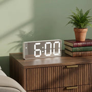 A modern digital alarm clock with a white rectangular body and glowing white LED numbers showing 6:00, placed on a wooden nightstand in a styled bedroom setting next to a potted fern and stacked books.