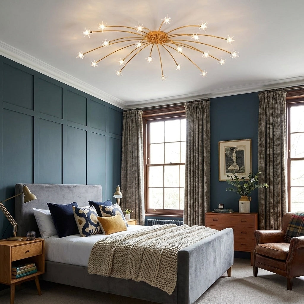 A modern multi-light chandelier with a gold starburst frame and glowing white spherical bulbs hangs in a styled bedroom with dark blue panelled walls, a grey bed, and warm wood furniture.