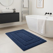 A dark blue rectangular bath mat with concentric raised borders lies on a light beige tiled bathroom floor next to a white freestanding bathtub with a white towel draped over the edge.