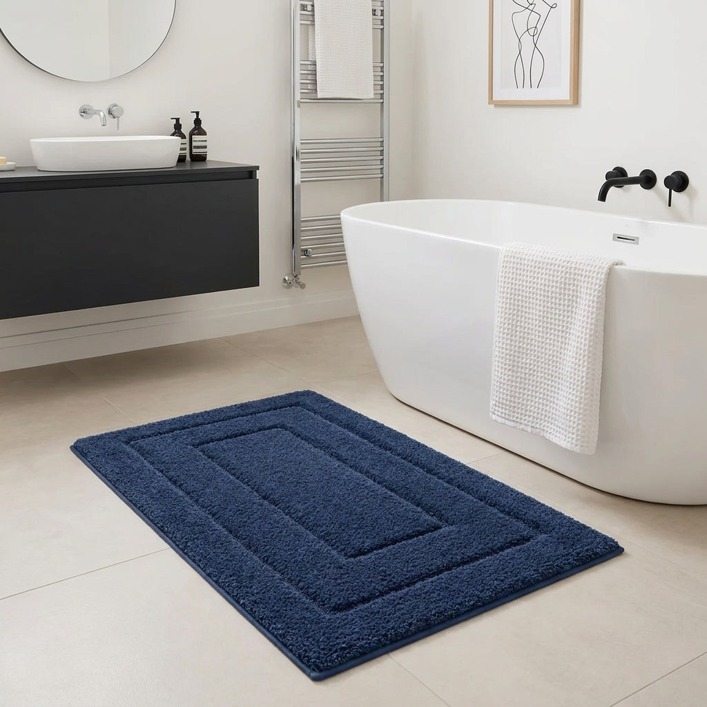 A dark blue rectangular bath mat with concentric raised borders lies on a light beige tiled bathroom floor next to a white freestanding bathtub with a white towel draped over the edge.