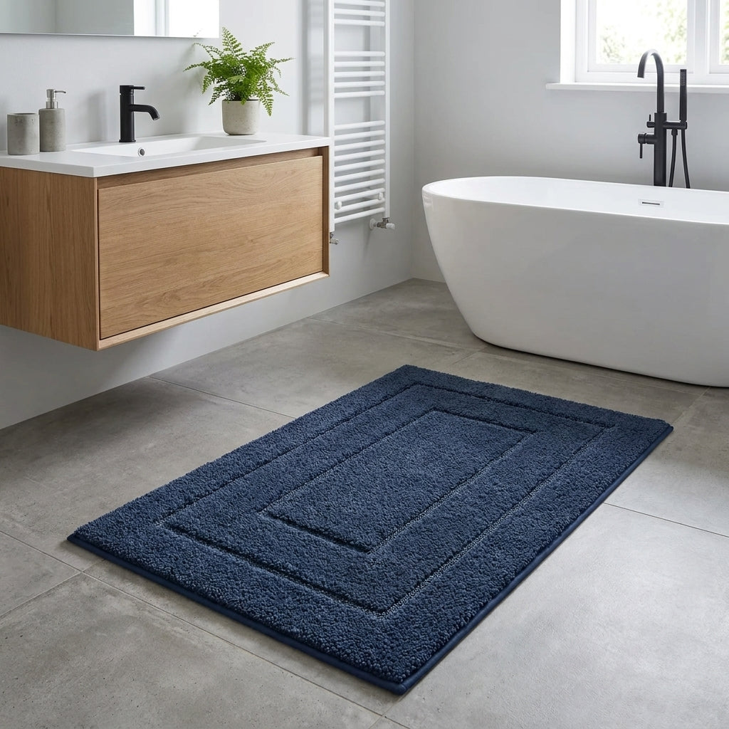 A dark navy blue rectangular bath mat with concentric square borders, shown on a light grey tiled bathroom floor next to a white freestanding bathtub and a floating wooden vanity.