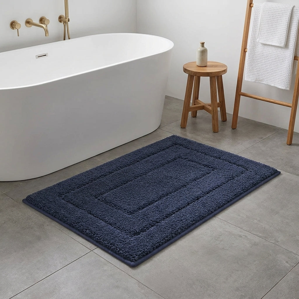 A dark navy blue rectangular bath mat with a raised geometric border design, shown on a grey tiled bathroom floor next to a white freestanding bathtub.