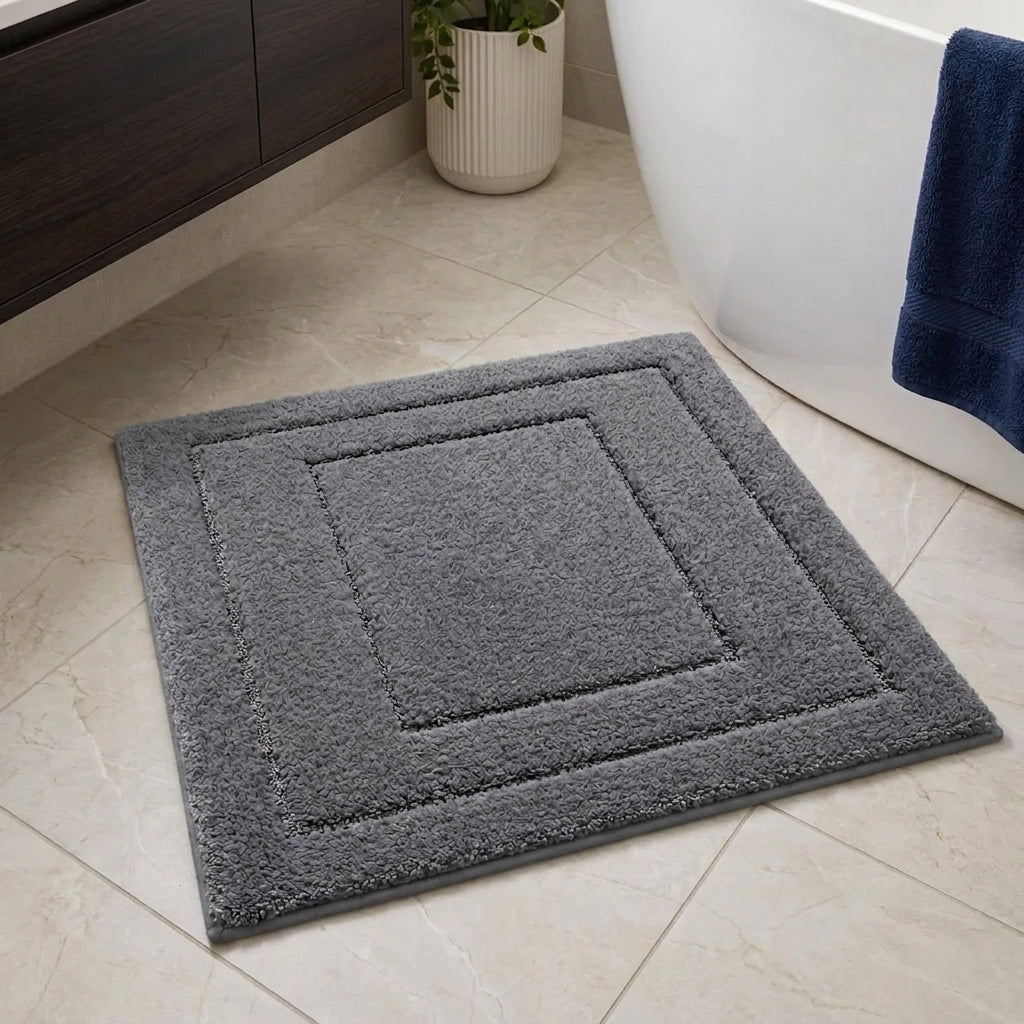 A square grey bath mat with concentric square borders and a plush, textured surface, shown on a beige tiled bathroom floor next to a white bathtub.