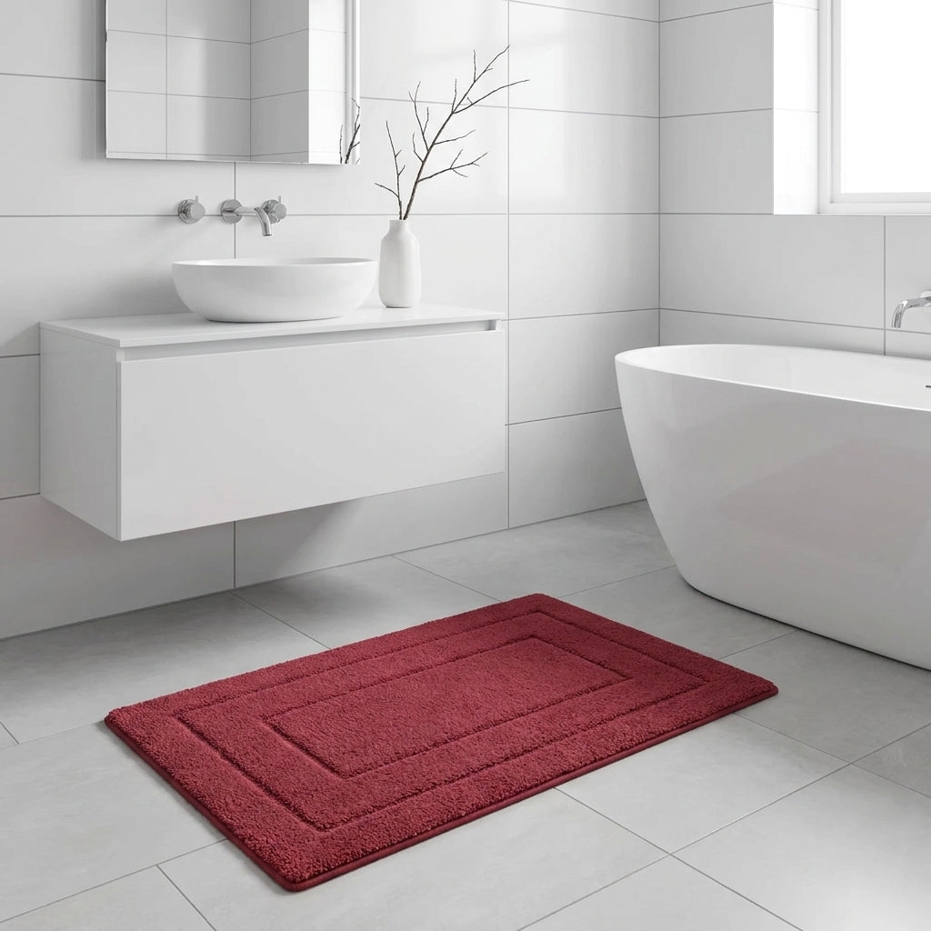 A rectangular red bath mat with concentric border stitching lies on a light grey tiled bathroom floor next to a white freestanding bathtub and a white wall-mounted sink.