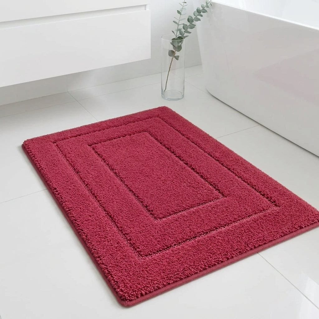 A rectangular red bath mat with concentric square borders, shown on a white tiled bathroom floor next to a bathtub.