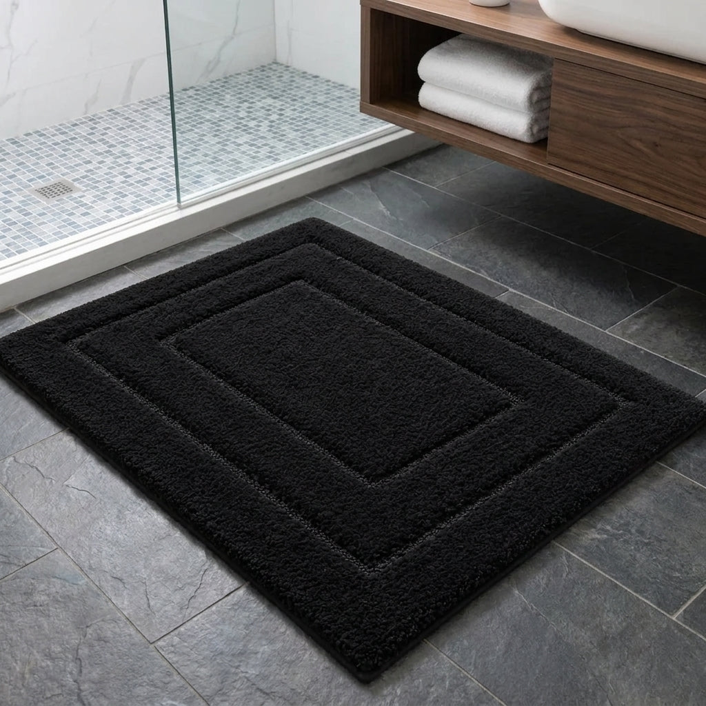 A rectangular black bath mat with concentric square borders and a shaggy texture, shown on a dark grey tiled bathroom floor next to a wooden vanity and glass shower enclosure.