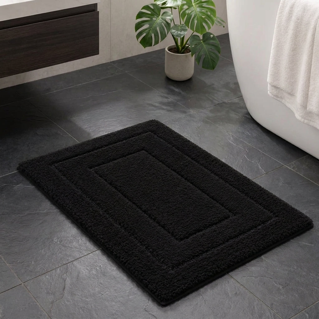 A black rectangular bath mat with concentric rectangular borders and a plush, textured surface, shown on a dark grey tiled bathroom floor next to a white bathtub with a white towel and a potted green plant.