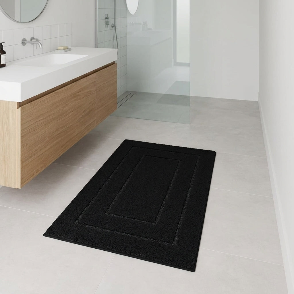 A black rectangular bath mat with concentric rectangular borders lies on a light grey tiled bathroom floor next to a floating wooden vanity with a white sink.