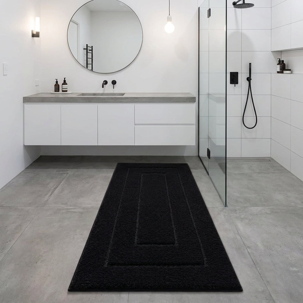 A long rectangular black bath mat with concentric rectangular cutouts lies on a grey tiled bathroom floor between a white floating vanity and a glass shower enclosure.