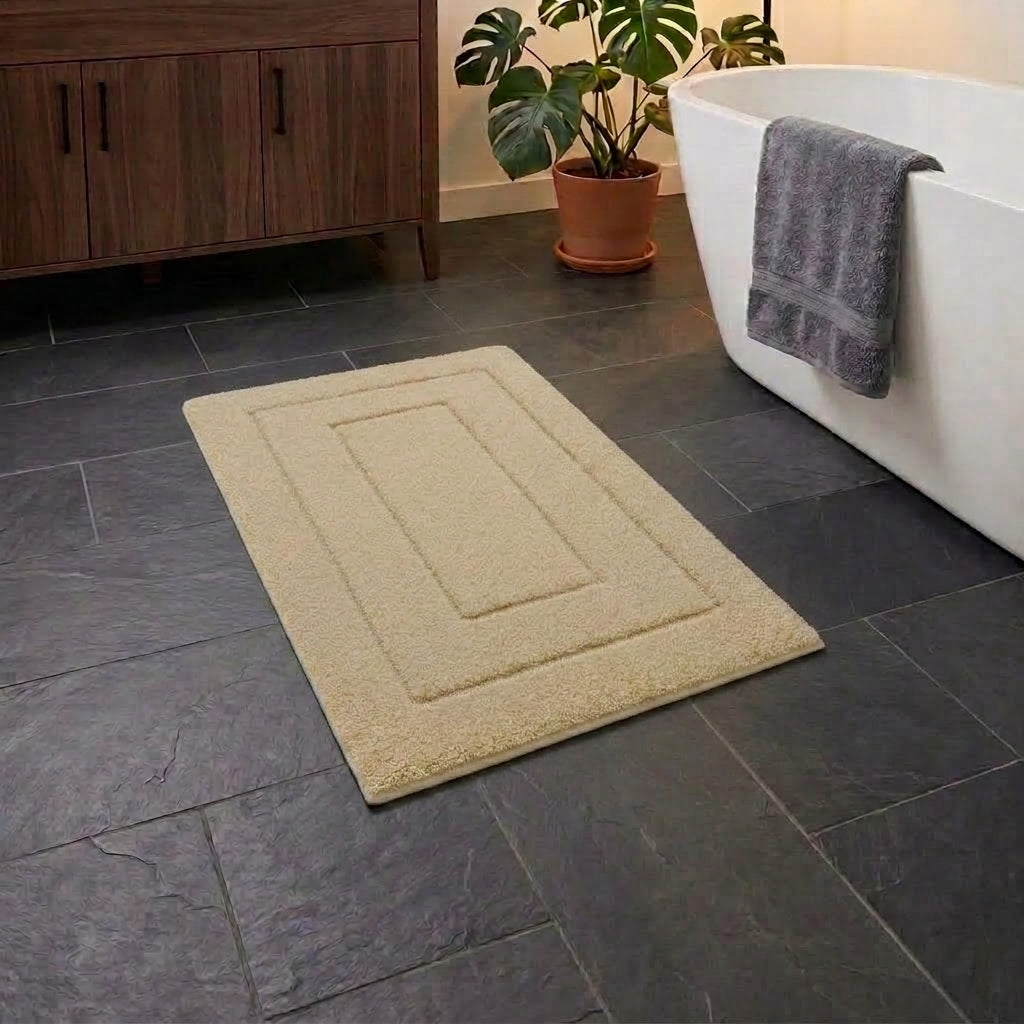 A rectangular beige bath mat with a raised border design, shown on a dark grey tiled bathroom floor next to a white bathtub with a grey towel and a potted plant.