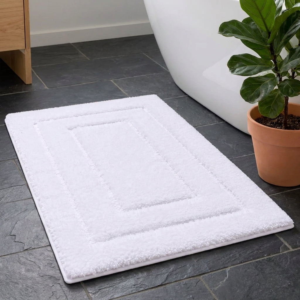 A white rectangular bath mat with a raised concentric border design, shown on a dark grey tiled bathroom floor next to a white bathtub and a potted fiddle-leaf fig plant.