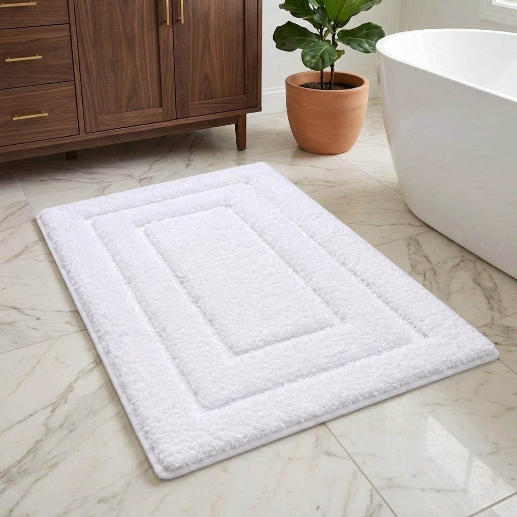A white rectangular bath mat with concentric rectangular borders and a plush, textured surface, shown on a light marble bathroom floor next to a white freestanding bathtub and a potted plant.