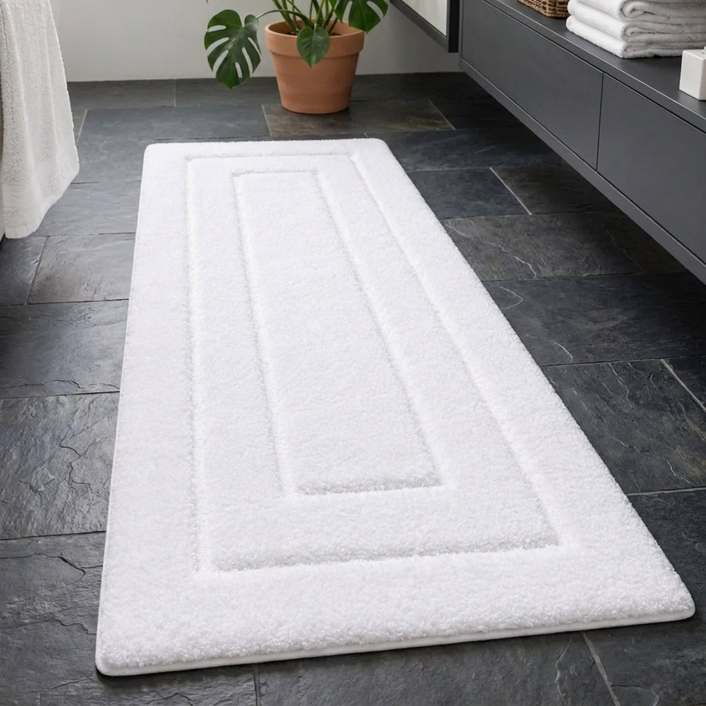 A white rectangular bath mat with a raised rectangular border design, shown on a dark slate tile floor next to a grey vanity cabinet with folded towels and a potted plant.