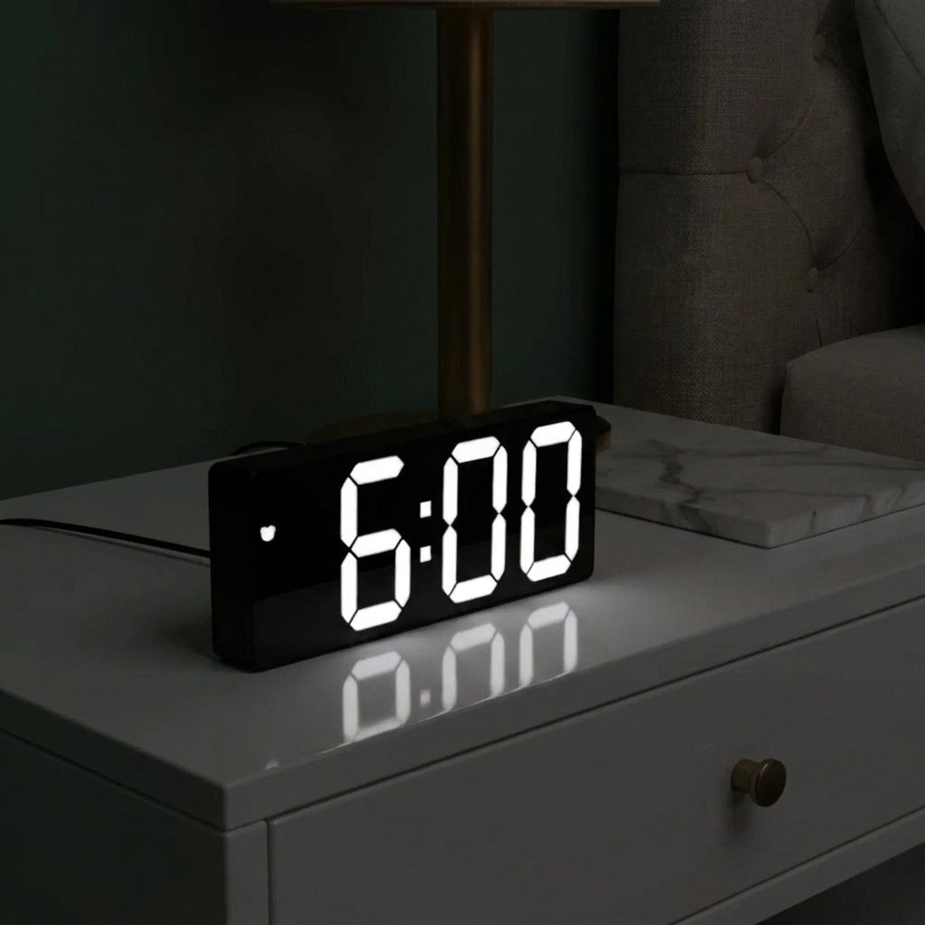 A black digital LED alarm clock displaying 6:00 in bright white digits, placed on a white bedside table in a dimly lit bedroom setting.