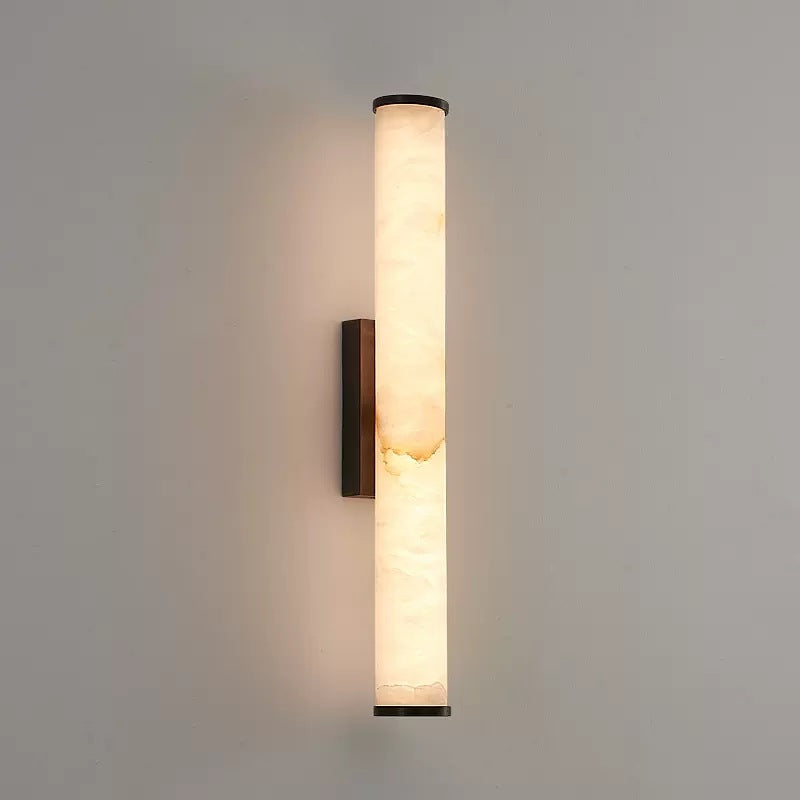 Vertical alabaster tube sconce with sleek black hardware glowing warmly on a plain neutral wall