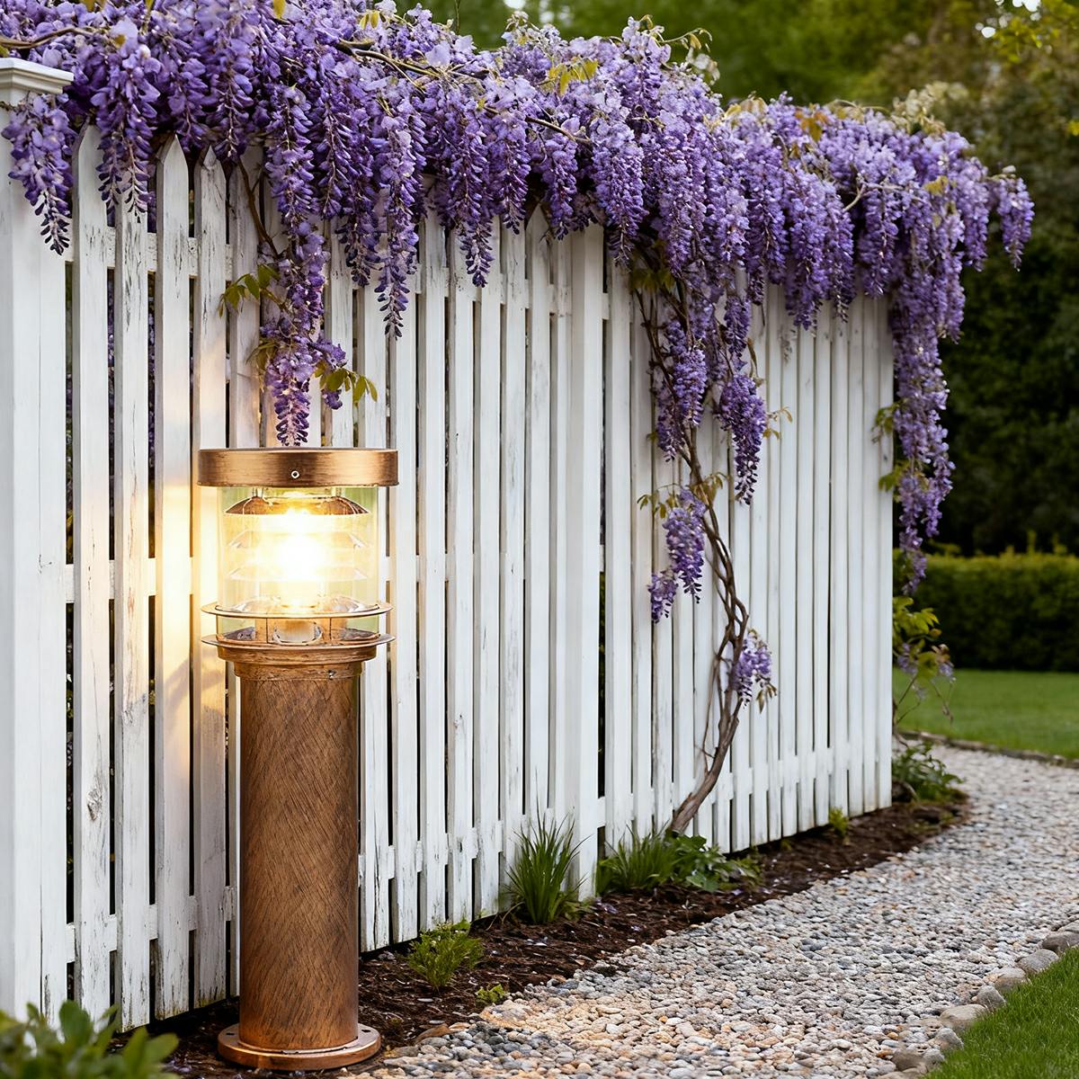 Bronze outdoor pillar light shining warmly next to a white garden fence and purple wisteria