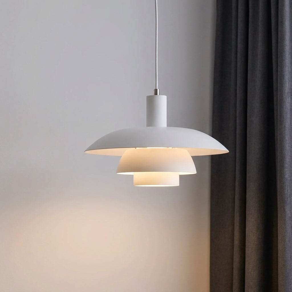 A modern white pendant light with a tiered, saucer-like design hangs from a cord against a light grey wall, casting a warm glow. A dark grey curtain is visible on the right side of the frame.