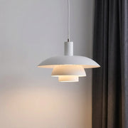 A modern white pendant light with a tiered, saucer-like design hangs from a cord against a light grey wall, casting a warm glow. A dark grey curtain is visible on the right side of the frame.