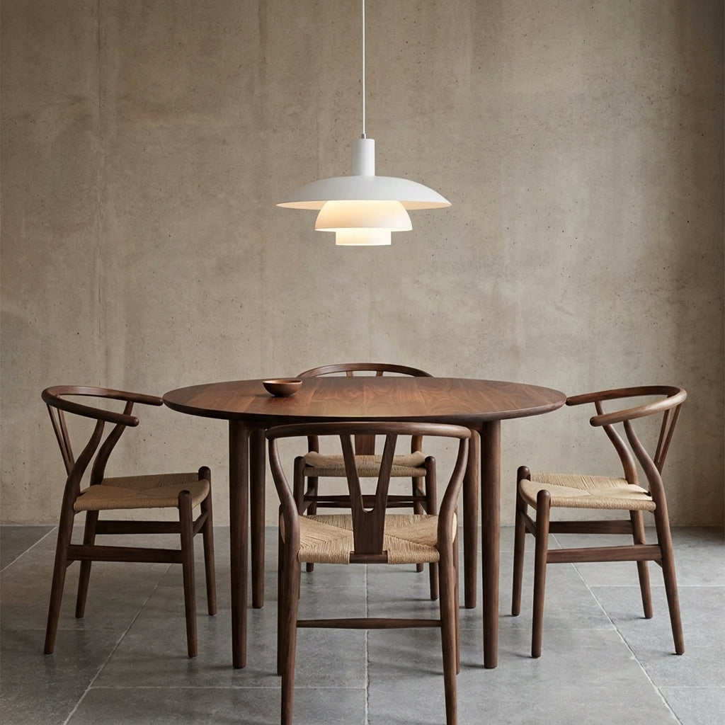A dining set featuring a round wooden table and four matching wooden chairs with woven seats, styled under a modern white tiered pendant light against a textured concrete wall.