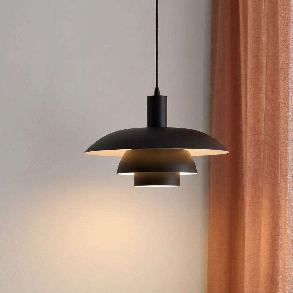 A modern black pendant light with a layered, disc-like shade hangs from a black cord against a light grey wall, casting a warm glow, with a rust-coloured curtain on the right.