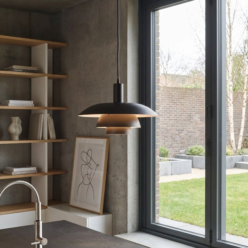 A modern black pendant light with a tiered, layered design hangs over a kitchen island in a contemporary kitchen. The room features a concrete wall, wooden floating shelves, and a large window looking out onto a brick garden wall.