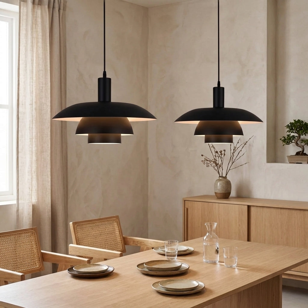 Two black pendant lights with a wide, shallow dome shade hang over a light wood dining table set with plates and glasses in a warm, modern dining room.