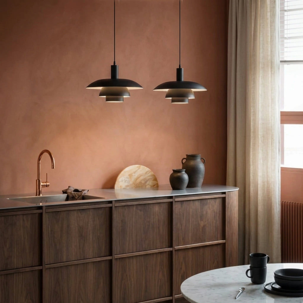 Two black metal pendant lights with tiered shades hang over a modern kitchen counter with dark wood cabinets, a copper faucet, and neutral-toned vases against a terracotta wall.