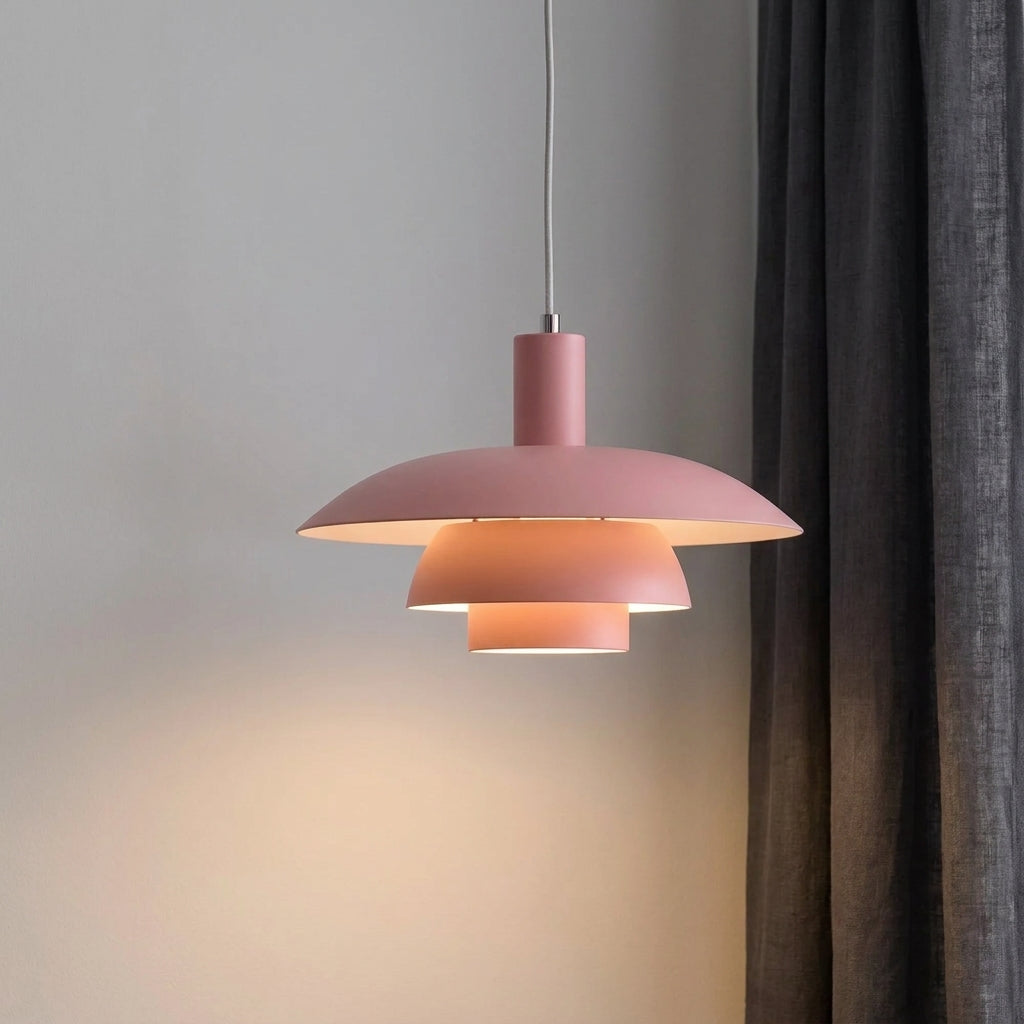 A modern tiered pendant light in pink colour, shown illuminated against a neutral grey wall next to a dark curtain.