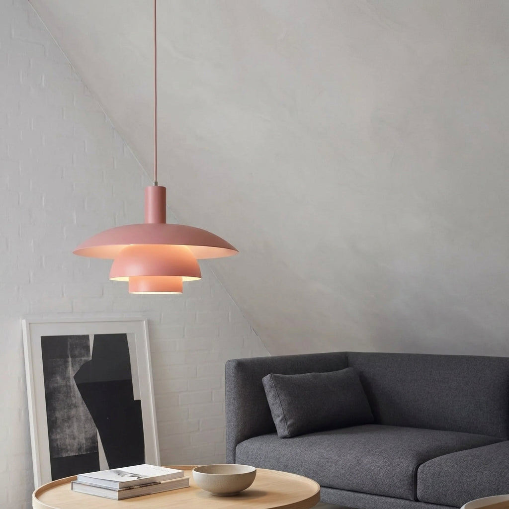 A modern tiered pendant light in a dusty rose pink colour, hanging in a minimalist living room setting with a grey sofa, round wooden table, and textured grey wall.