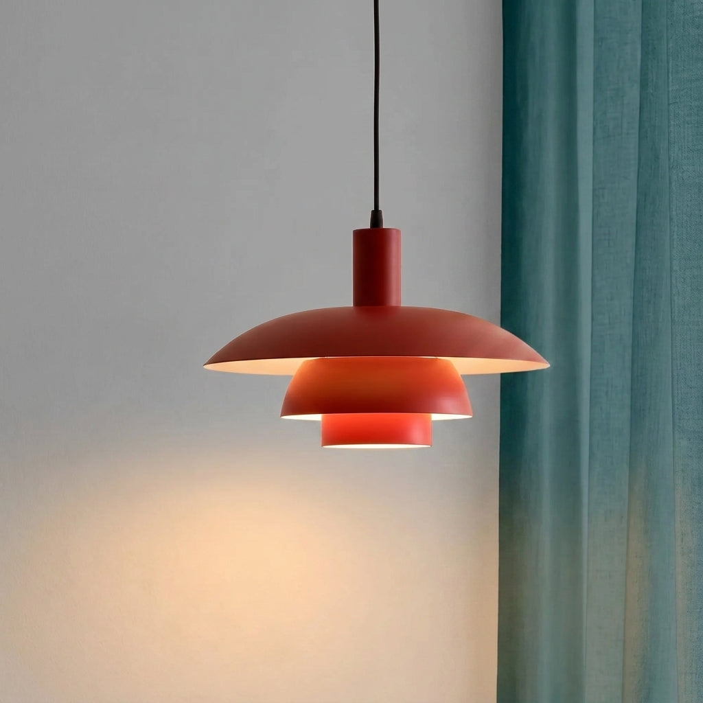 A modern tiered pendant light with a matte red finish, suspended by a black cord against a neutral grey wall next to a teal curtain.