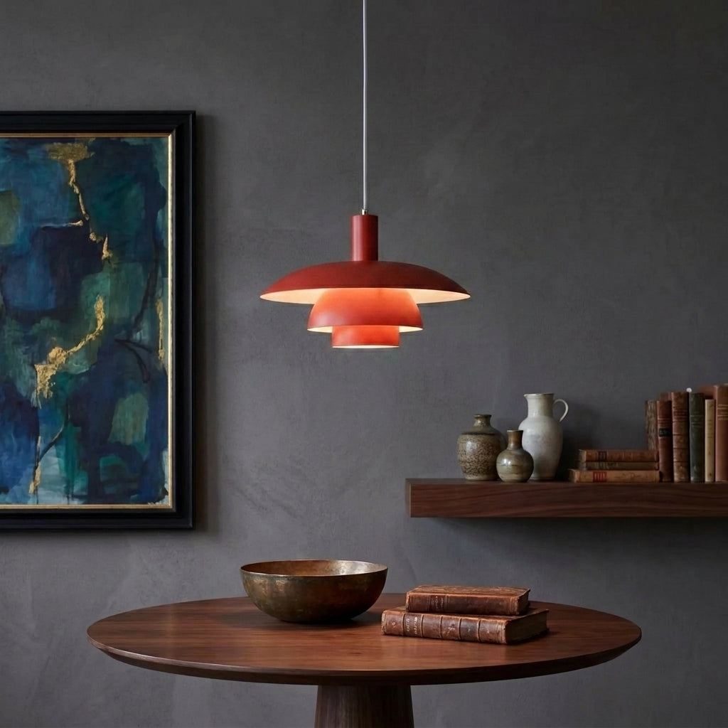 A modern tiered pendant light with a red shade hangs over a round wooden table in a styled room with a dark grey textured wall, framed abstract art, and a floating shelf with books and vases.