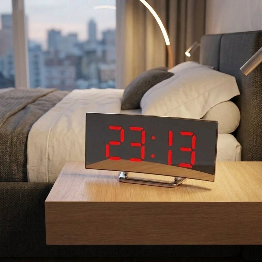 A digital alarm clock with a black face and red LED numbers sits on a wooden bedside table in a modern bedroom setting.