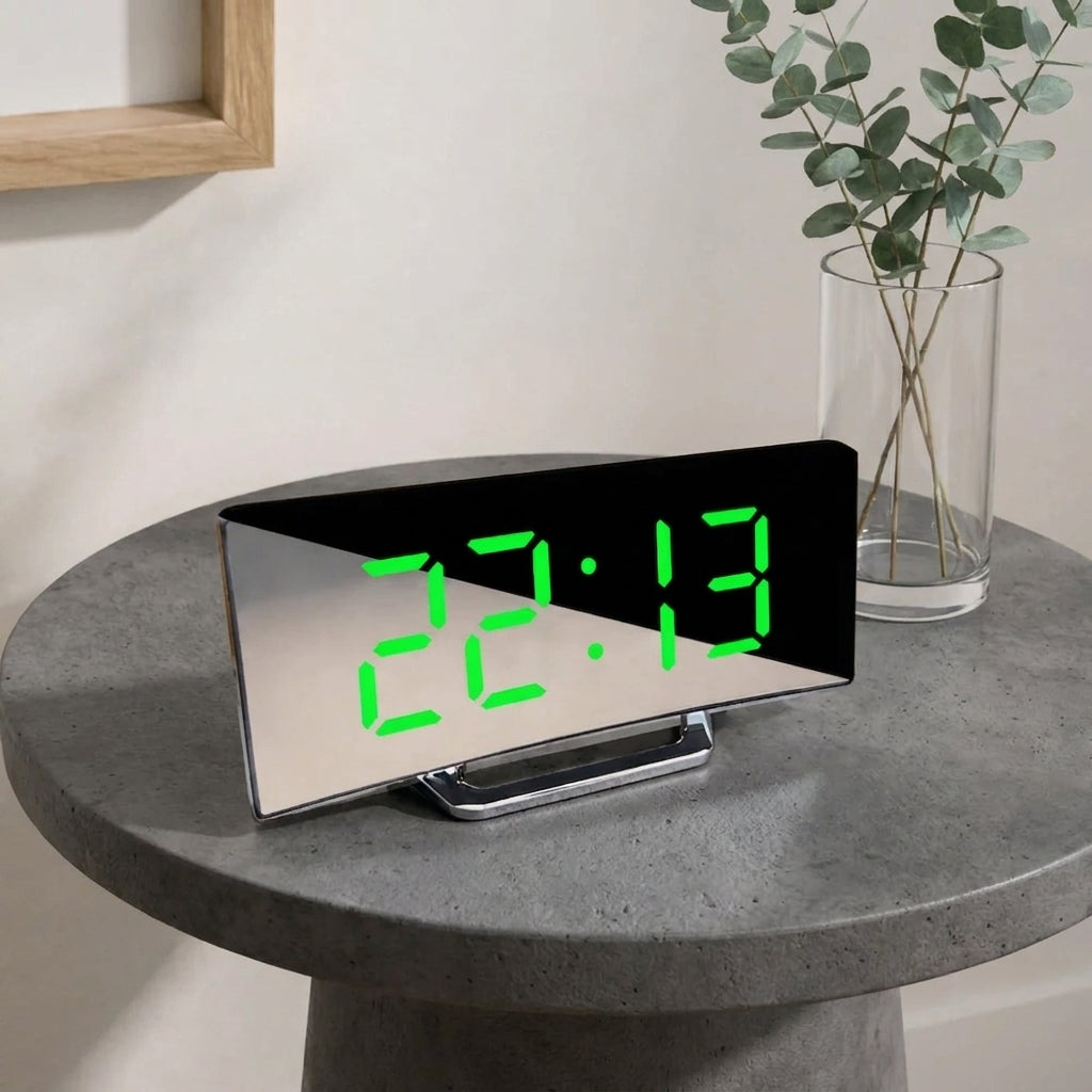 A modern digital alarm clock with a black mirrored rectangular face and bright green LED numbers, displayed on a round grey stone table next to a glass vase with eucalyptus branches.