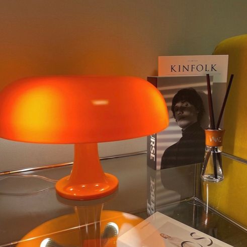 Glossy orange retro mushroom lamp glowing warmly on a glass shelf next to a Kinfolk book