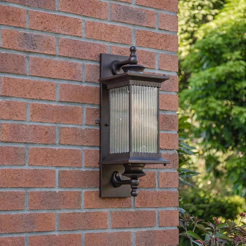 A brown outdoor wall sconce with a ribbed glass shade, mounted on a red brick wall with green foliage visible on the right.