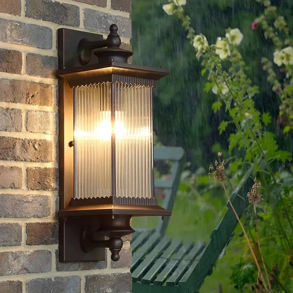 An outdoor wall-mounted lantern light fixture with a warm glowing bulb, featuring a dark brown metal frame and ribbed clear glass panels, mounted on a brick wall with a garden background visible in the rain.
