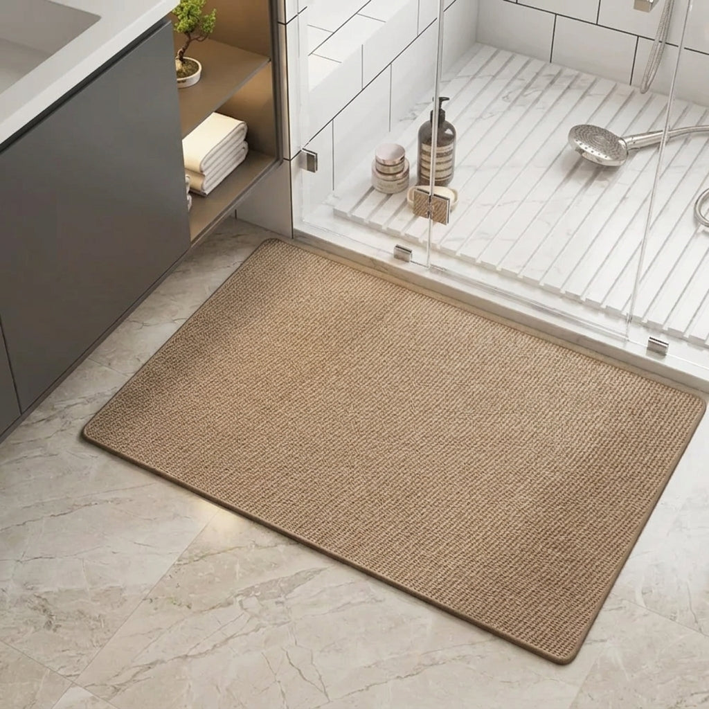 A rectangular woven bath mat in a brown colour, shown on a light marble bathroom floor next to a glass shower door.
