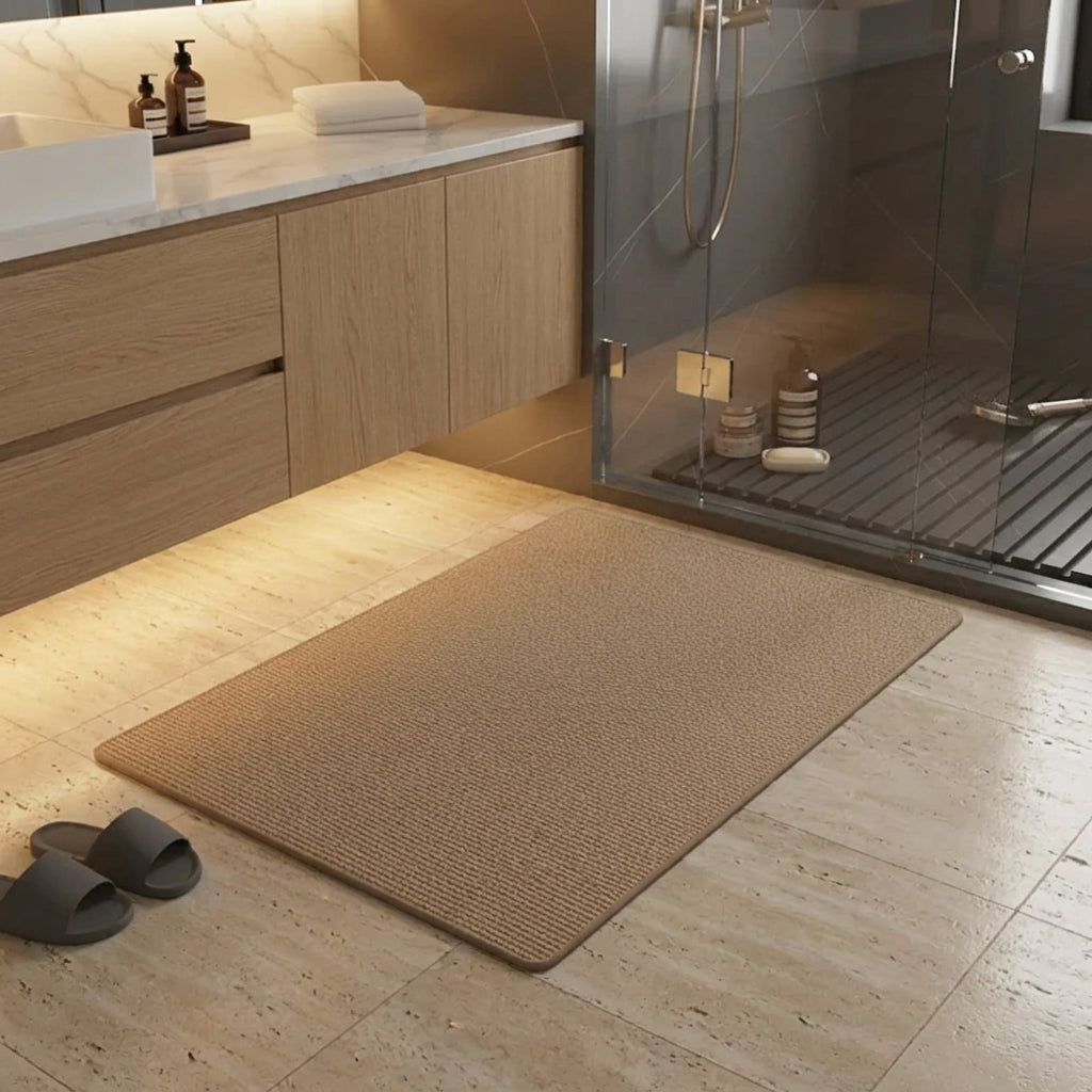 A rectangular brown bath mat with a textured woven surface lies on a light stone bathroom floor. A glass shower enclosure is visible in the background, along with a vanity, toiletries, and a pair of dark grey slide sandals nearby.