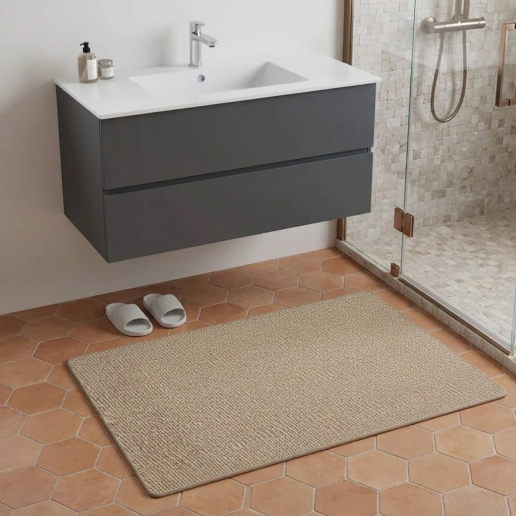 A rectangular brown woven bath mat on a terracotta hexagonal tile floor in a modern bathroom, positioned in front of a floating dark grey vanity with a white sink and next to a glass shower enclosure.