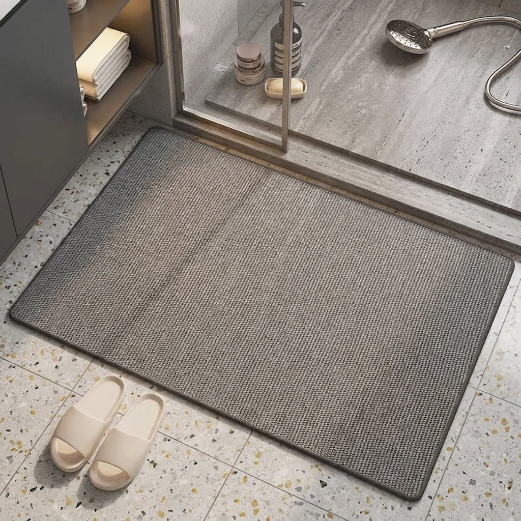 A rectangular grey textured bath mat placed on a speckled bathroom floor near a glass shower door, with a pair of off-white slippers nearby.