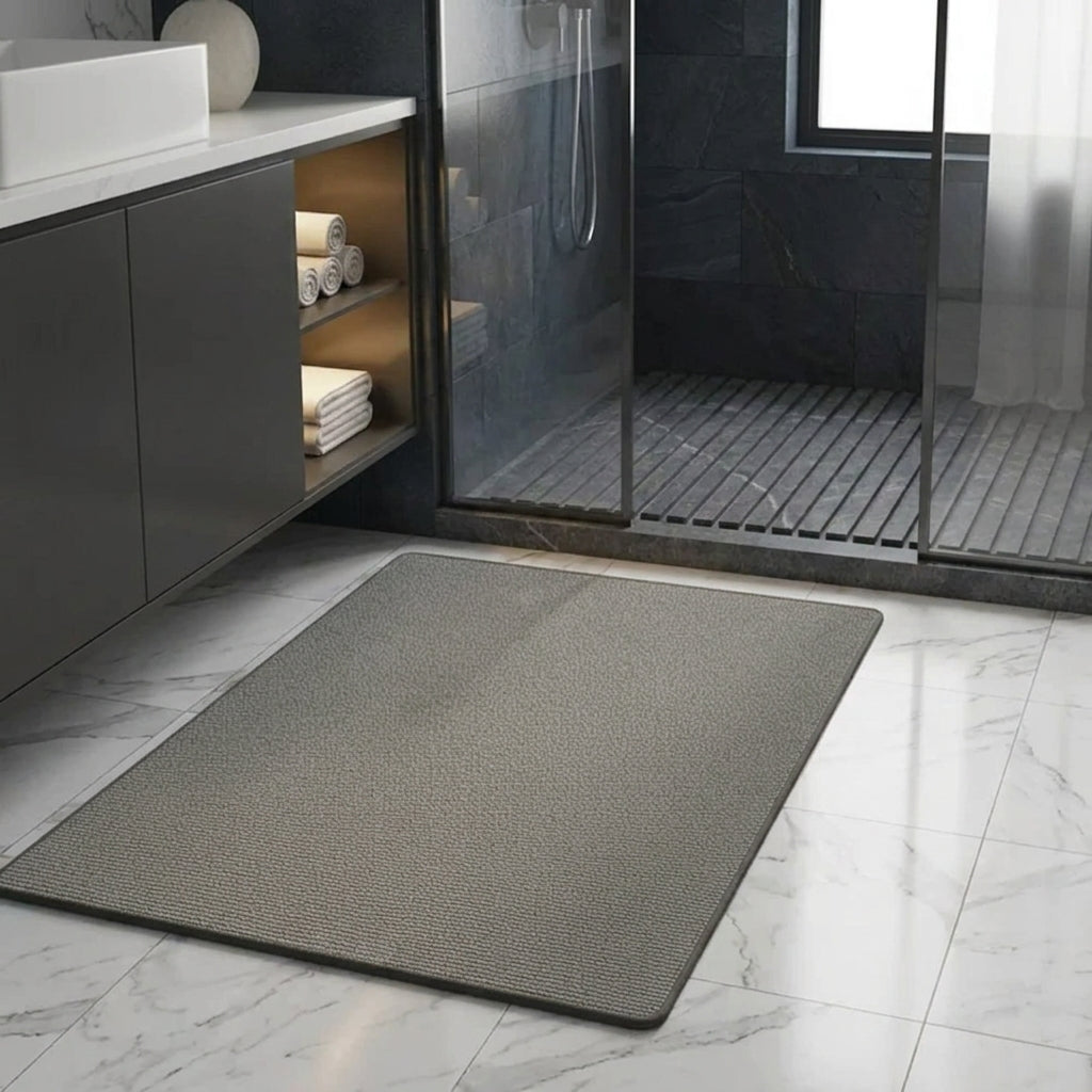 A rectangular grey bath mat with a textured surface lies on a white marble-look bathroom floor in front of a glass shower enclosure and dark vanity cabinet.