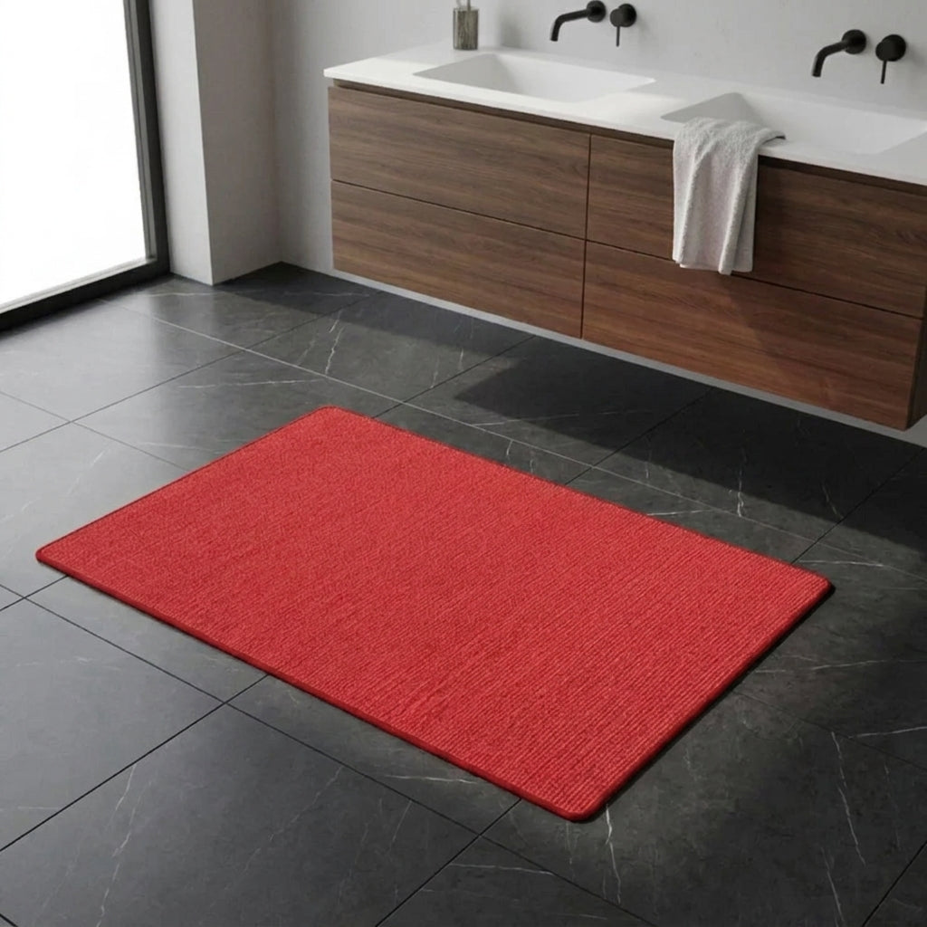 A bright red rectangular bath mat placed on a dark grey tiled bathroom floor in front of a floating wooden vanity with a white sink and a white towel.