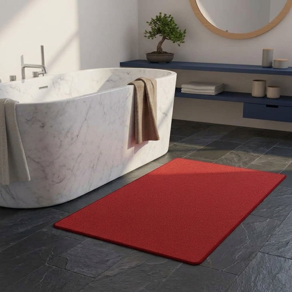 A rectangular red bath mat placed on a dark slate tile floor next to a white marble freestanding bathtub in a modern bathroom setting.