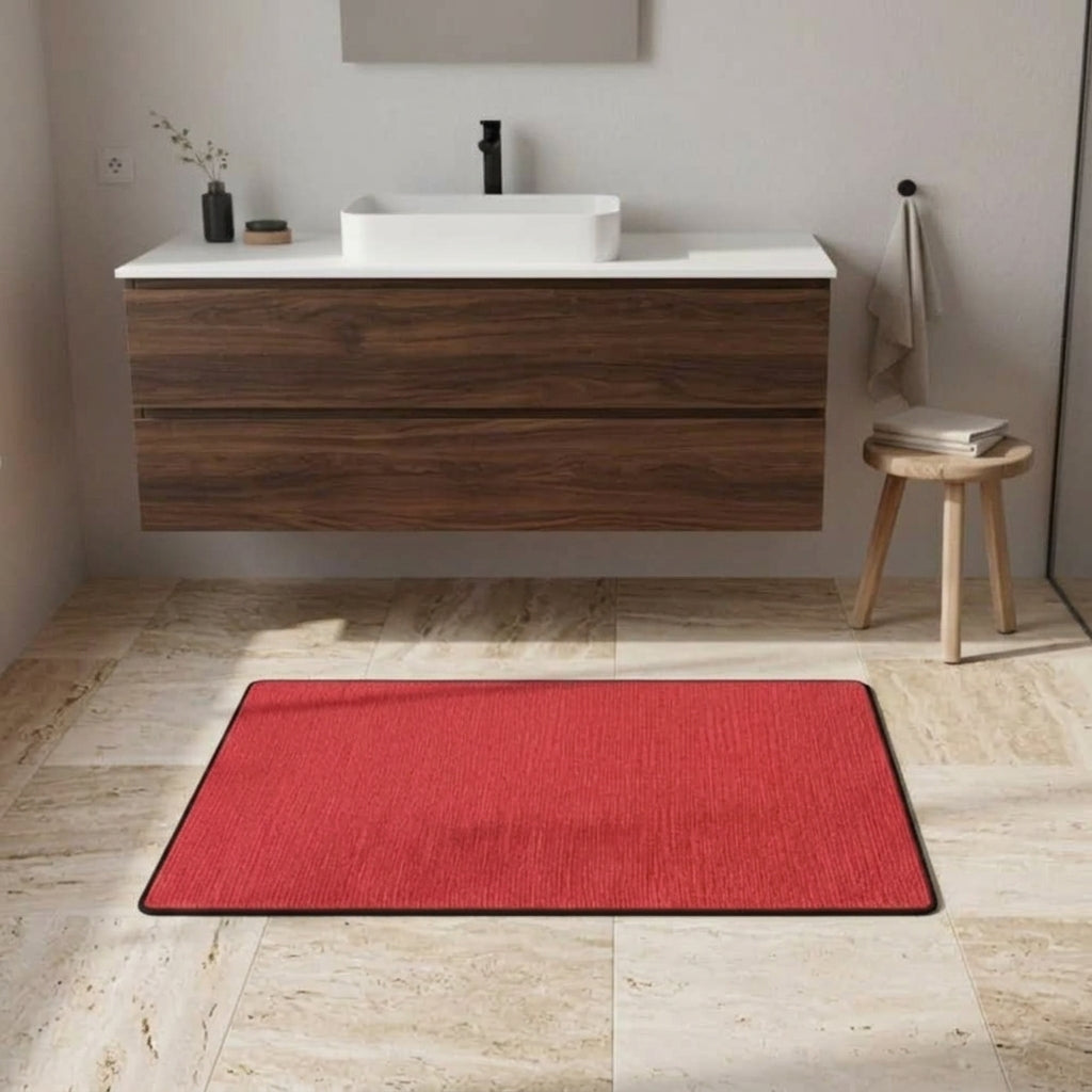 A rectangular red bath mat with a black border lies on a light beige stone-tiled bathroom floor in front of a floating dark wood vanity with a white sink and countertop.