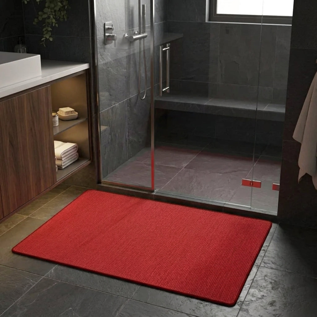A rectangular red bath mat with a textured, ribbed surface lies on a dark grey tiled bathroom floor in front of a glass shower enclosure.