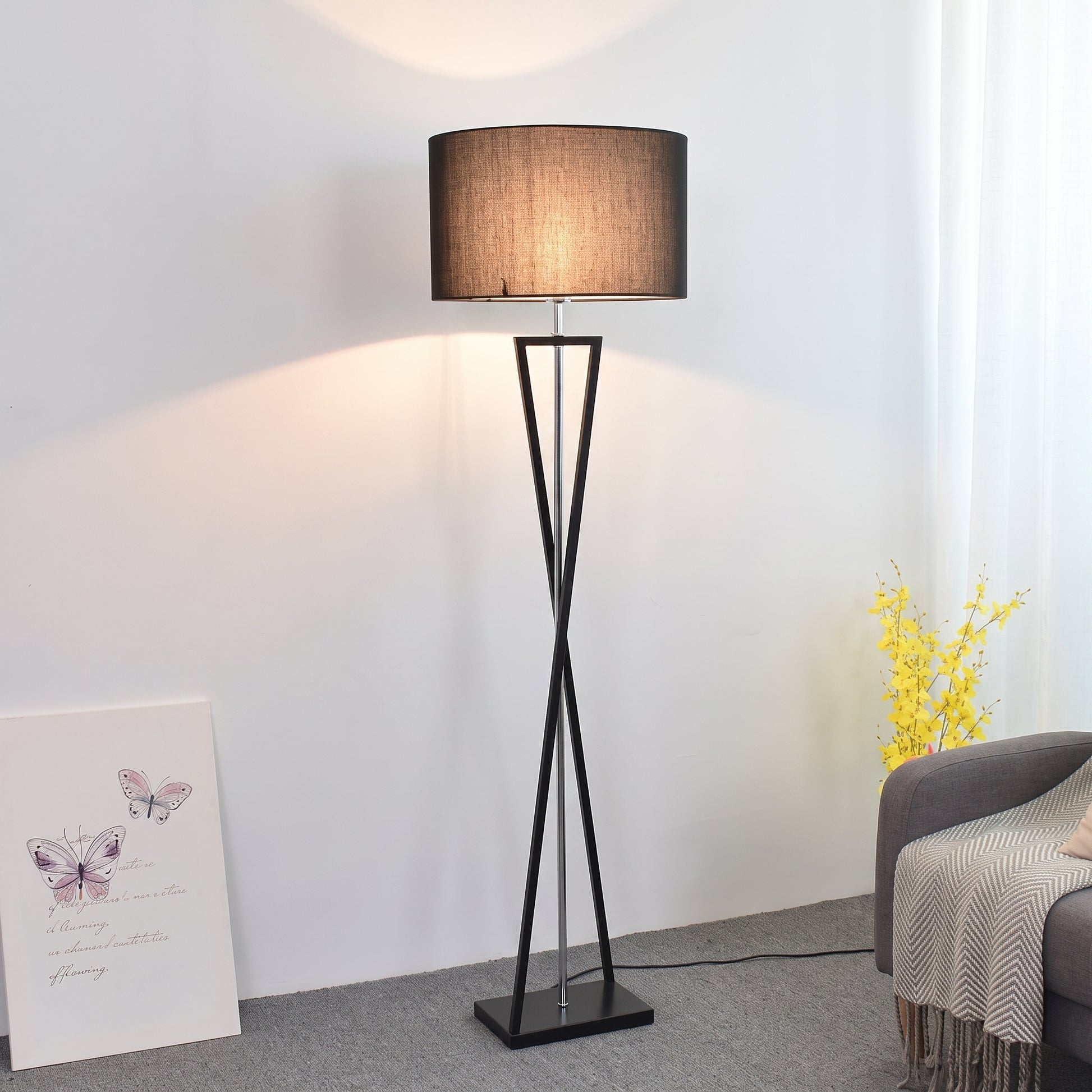 A modern mopij floor lamp featuring an intersecting matte black iron frame in an hourglass silhouette, topped with a deep black fabric drum shade emitting a warm ambient glow, positioned in a bright, contemporary living space next to a grey sofa