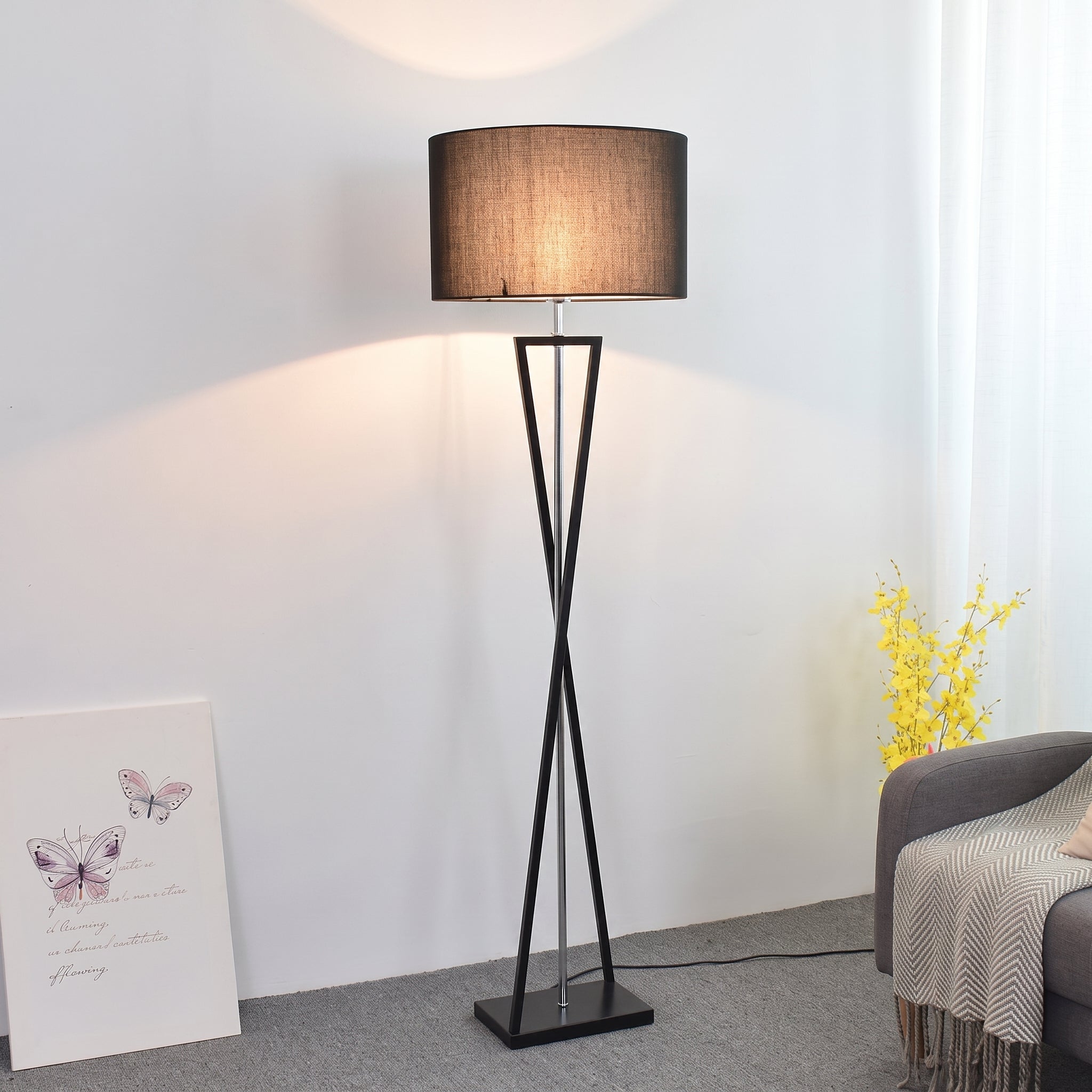 A modern mopij floor lamp featuring an intersecting matte black iron frame in an hourglass silhouette, topped with a deep black fabric drum shade emitting a warm ambient glow, positioned in a bright, contemporary living space next to a grey sofa