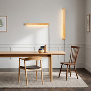 Nizku slim linear pine wood wall lamps mounted on a wall beside a wooden dining table and chairs, with a plant and framed art