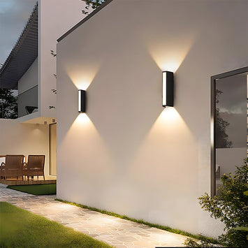Two modern black linear peqi LED outdoor wall lights casting up and down illumination on a white house exterior